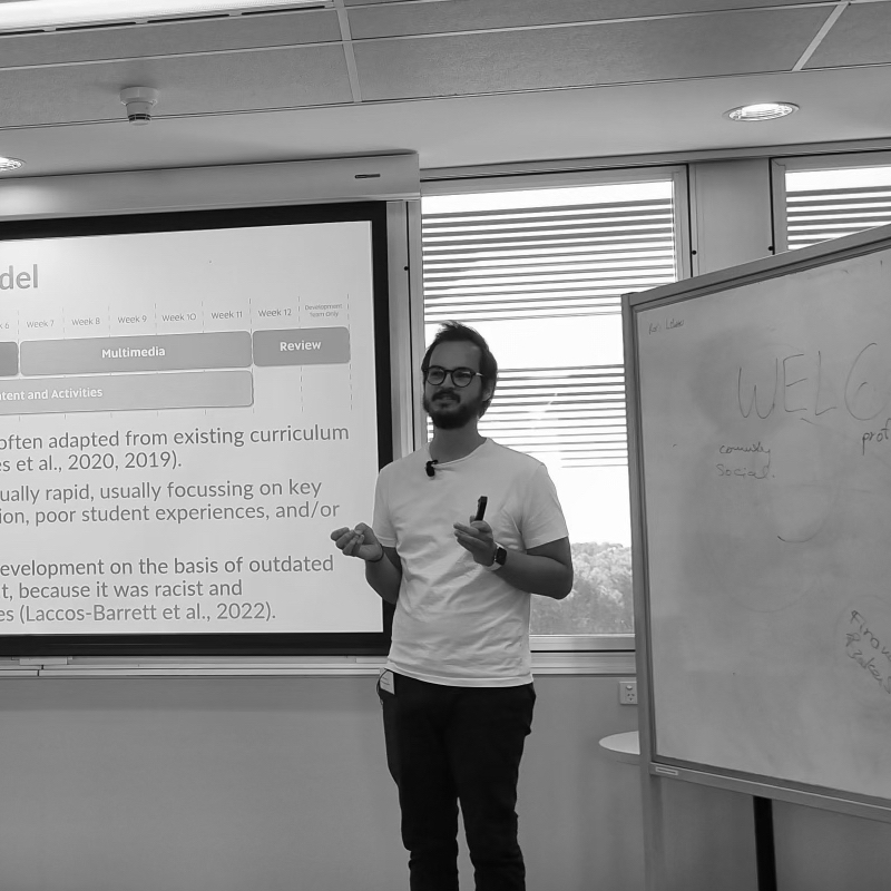 A human standing at the front of a lecture theatre wearing a white t-shirt, moving arms animatedly. Backdrop including a blank whiteboard and a presentation screen showing a slide.
