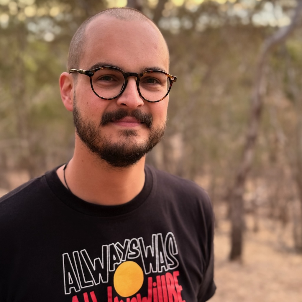 A semi-professional photo of a human head and shoulders, none hair, glasses, beard, looking toward you with a smile.